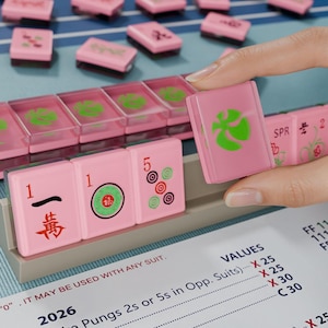 A hand is holding a pink tile with a green design, while a table with pink tiles and a blue surface is visible in the background.