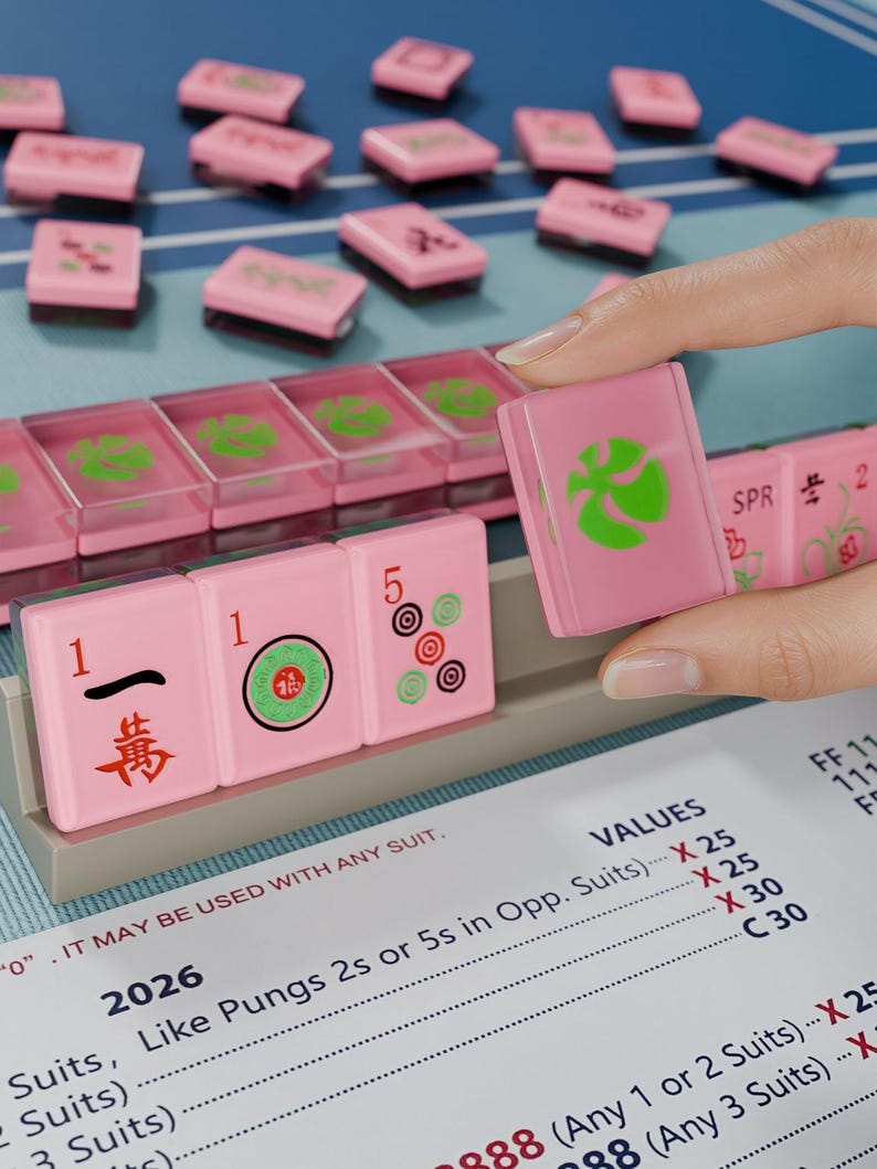 A hand is holding a pink tile with a green design, while a table with pink tiles and a blue surface is visible in the background.