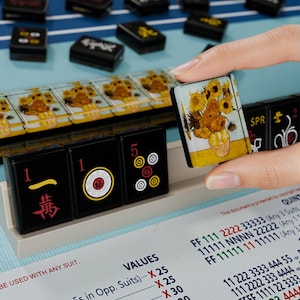 A hand is holding a small square-shaped object with a picture of flowers on it, in front of a collection of black and yellow tiles or blocks with various designs and numbers on them.