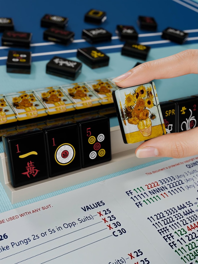 A hand is holding a small square-shaped object with a picture of flowers on it, in front of a collection of black and yellow tiles or blocks with various designs and numbers on them.