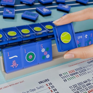 A hand is holding a blue tile with a tennis ball design, and there are several blue tiles with tennis ball designs on a table.