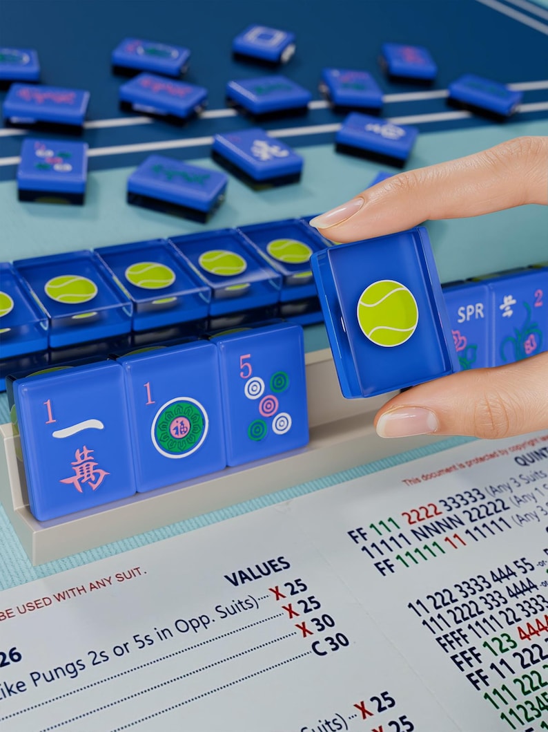 A hand is holding a blue tile with a tennis ball design, and there are several blue tiles with tennis ball designs on a table.
