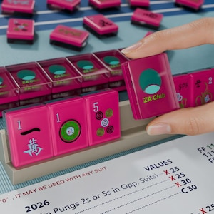 A hand is holding a pink tile with a logo, while a table with a list of items is visible in the background.