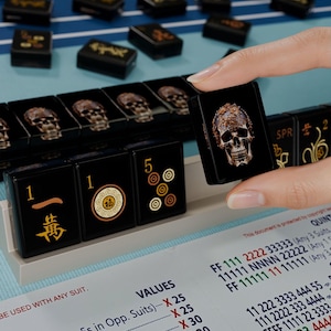 A hand is holding a black game piece with a skull design, while a table with a blue surface displays a list of game pieces and their values.