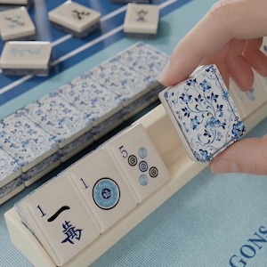 A hand is holding a blue and white tile with a floral pattern, which appears to be a game piece for mahjong. The tiles are arranged on a blue and white surface, and there are additional tiles visible in the background.