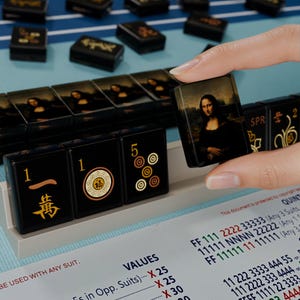 A hand is holding a small square with a picture of the Mona Lisa on it, in front of a collection of black and gold dominoes.
