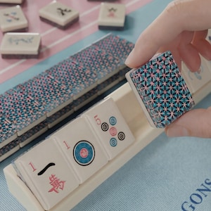 A hand is holding a tile with a geometric pattern, while a set of dominoes is visible in the background.