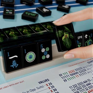 A hand is holding a black tile with a green leaf design, surrounded by a collection of black tiles with various designs and numbers on them, all placed on a blue surface.