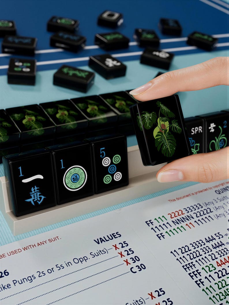 A hand is holding a black tile with a green leaf design, surrounded by a collection of black tiles with various designs and numbers on them, all placed on a blue surface.