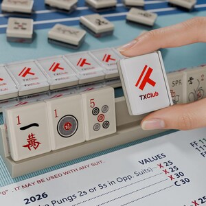 A hand is holding a tile with the TX Club logo, while a game of mahjong is visible in the background.