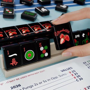 a hand holding a black tile with a strawberry design, surrounded by a collection of black tiles with red and green designs, all placed on a blue surface.