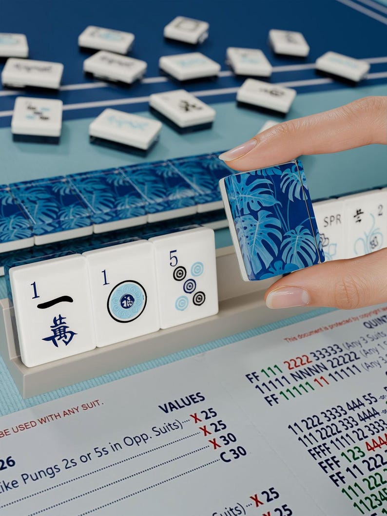 A hand is holding a blue and white tile with a floral pattern, while a table with a blue and white tablecloth and a blue and white tile display is visible in the background.
