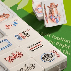 May include: A close-up of a Mahjong set with white tiles. Some tiles have red and black symbols, while others feature a horse wearing a cowboy hat and a bandana. The word "JOKER" is visible on one tile.