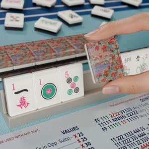 A hand is holding a small, colorful tile in front of a table with a blue surface and a variety of tiles and blocks on it.
