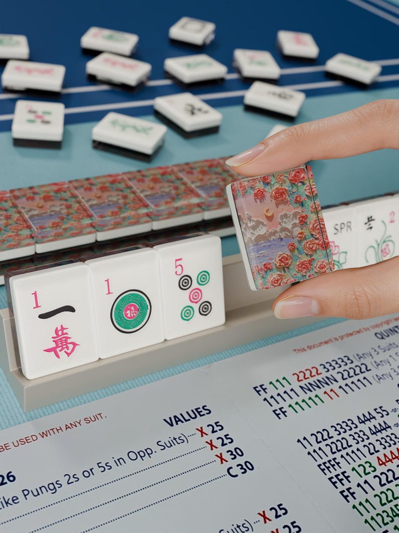 A hand is holding a small, colorful tile in front of a table with a blue surface and a variety of tiles and blocks on it.