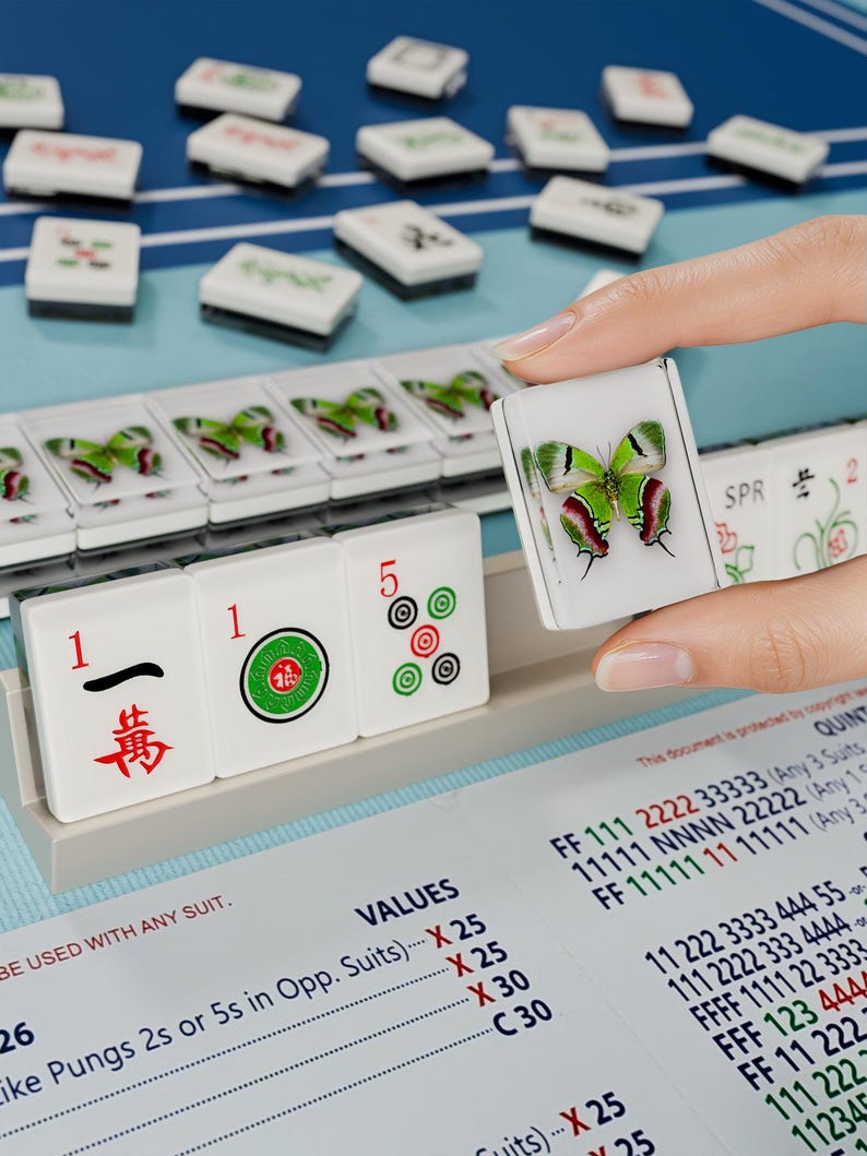 A hand is holding a tile with a butterfly design, while a table with a blue surface displays a collection of tiles and a price list.