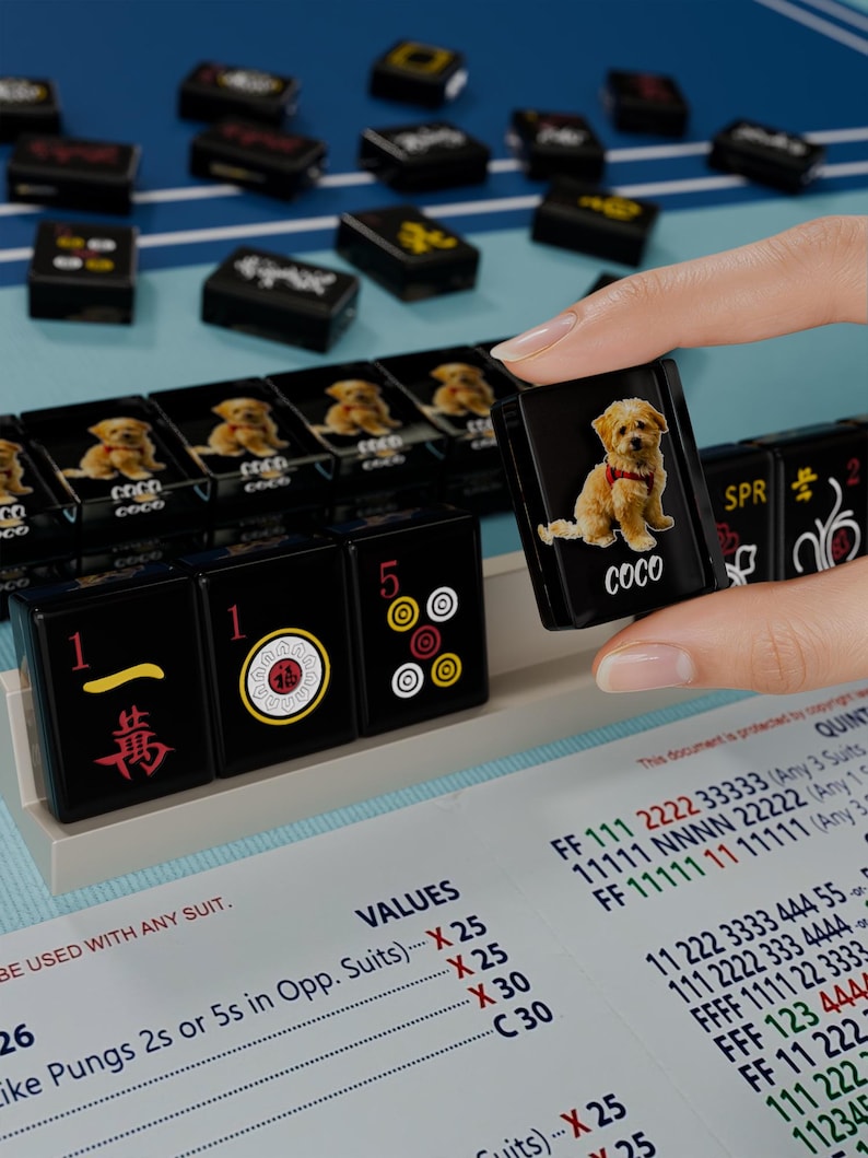 A hand is holding a small black game piece with a picture of a dog on it, in front of a collection of black and yellow game pieces on a blue surface.