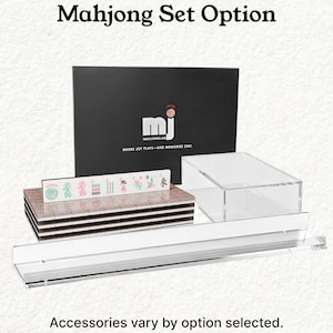 a set of mahjong tiles and accessories, including a black box with the mahjong logo and text, a stack of tiles, and a clear glass display case.