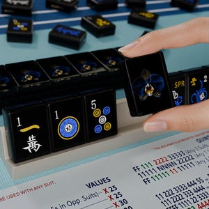 A hand is holding a black game piece with a blue and yellow design, standing in front of a table with a blue and white checkered pattern.