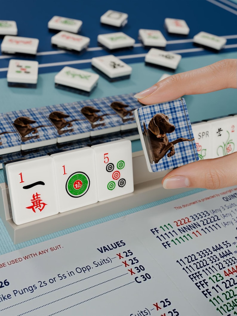 A hand is holding a tile with a dog image, while a game of mahjong is being played on a table with tiles and a score sheet.