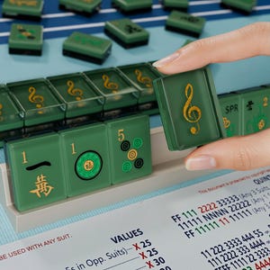 A hand is holding a green tile with a musical note symbol on it, with a background of green tiles and a sheet of paper with numbers and text visible.