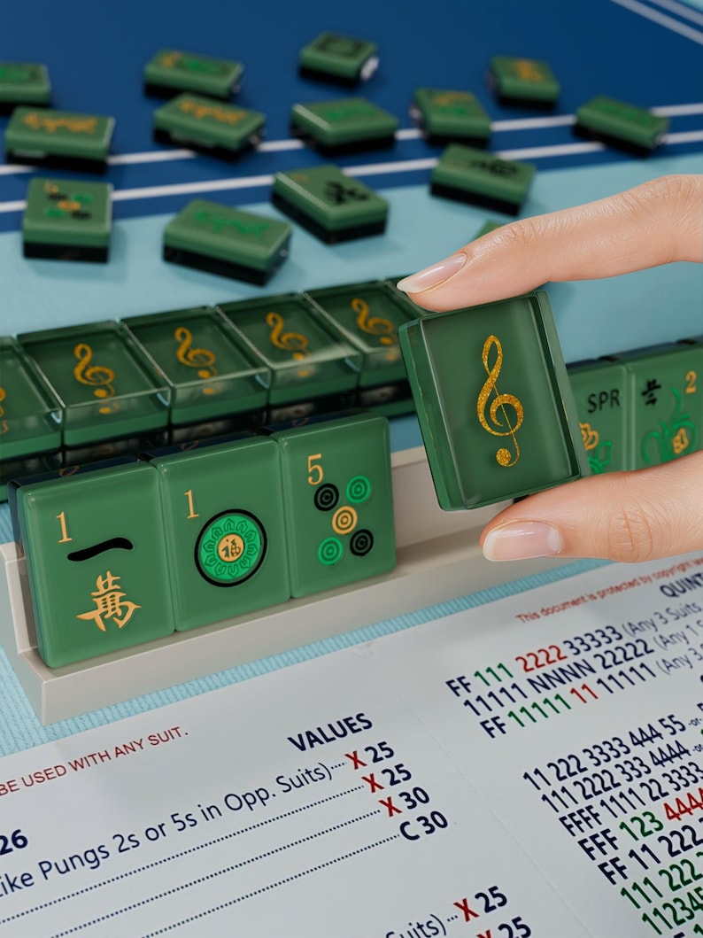 A hand is holding a green tile with a musical note symbol on it, with a background of green tiles and a sheet of paper with numbers and text visible.