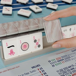A hand is holding a marble tile in front of a table with a game board and tiles, suggesting a game of mahjong or similar.