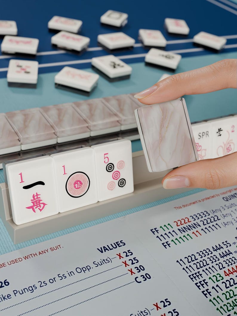 A hand is holding a marble tile in front of a table with a game board and tiles, suggesting a game of mahjong or similar.