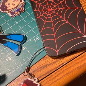 Puede incluir: Funda de tel&eacute;fono negra con un dise&ntilde;o de telara&ntilde;a roja. Un peque&ntilde;o dije con un personaje de dibujos animados est&aacute; adjunto. Tambi&eacute;n son visibles otros dijes de personajes de dibujos animados. Los art&iacute;culos est&aacute;n sobre una esterilla de corte verde y una superficie de madera.