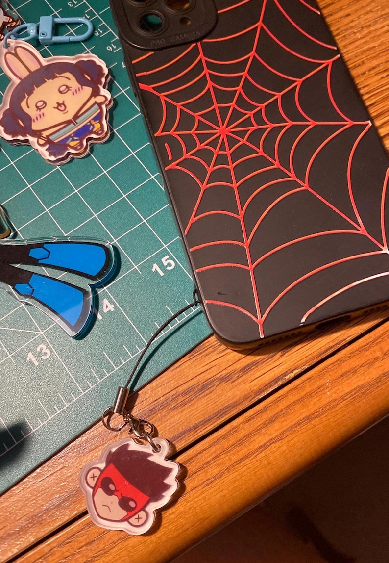 Puede incluir: Funda de tel&eacute;fono negra con un dise&ntilde;o de telara&ntilde;a roja. Un peque&ntilde;o dije con un personaje de dibujos animados est&aacute; adjunto. Tambi&eacute;n son visibles otros dijes de personajes de dibujos animados. Los art&iacute;culos est&aacute;n sobre una esterilla de corte verde y una superficie de madera.