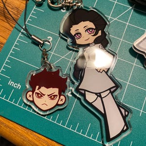 Puede incluir: Dos colgantes acr&iacute;licos de estilo anime con herrajes plateados. Un colgante presenta un personaje pelirrojo con expresi&oacute;n enfadada. El otro colgante representa a una mujer con un atuendo gris y ojos rosas.