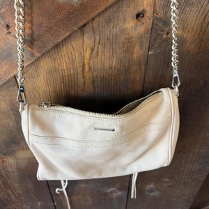 Peut inclure: Un sac &agrave; main cylindrique cr&egrave;me avec une bandouli&egrave;re en cha&icirc;ne argent&eacute;e. Le sac a une fermeture &eacute;clair et un petit logo en m&eacute;tal. Deux glands cr&egrave;me pendent du bas du sac. Le sac est suspendu sur un fond en bois.