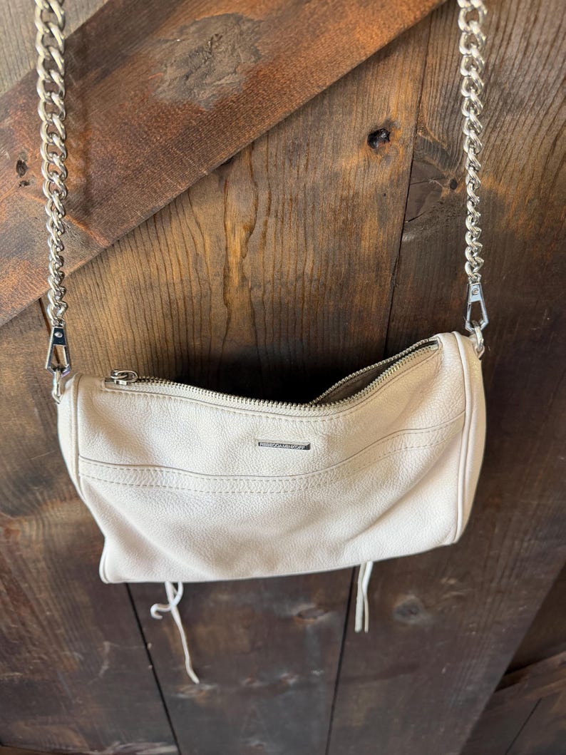 Peut inclure: Un sac &agrave; main cylindrique cr&egrave;me avec une bandouli&egrave;re en cha&icirc;ne argent&eacute;e. Le sac a une fermeture &eacute;clair et un petit logo en m&eacute;tal. Deux glands cr&egrave;me pendent du bas du sac. Le sac est suspendu sur un fond en bois.