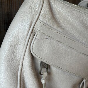 Peut inclure: Gros plan sur un sac &agrave; main en cuir gris clair. Le sac est dot&eacute; d'une fermeture &eacute;clair argent&eacute;e et d'une petite poche. Le cuir a un aspect textur&eacute; avec des coutures visibles et une bandouli&egrave;re en cha&icirc;ne argent&eacute;e.