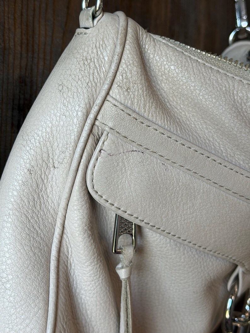 Peut inclure: Gros plan sur un sac &agrave; main en cuir gris clair. Le sac est dot&eacute; d'une fermeture &eacute;clair argent&eacute;e et d'une petite poche. Le cuir a un aspect textur&eacute; avec des coutures visibles et une bandouli&egrave;re en cha&icirc;ne argent&eacute;e.