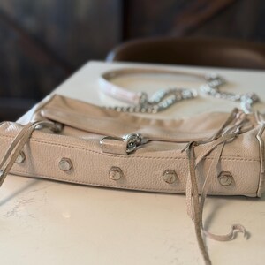 Peut inclure: Un sac &agrave; main en cuir rose clair avec des &eacute;l&eacute;ments m&eacute;talliques argent&eacute;s, dont des clous et une bandouli&egrave;re en cha&icirc;ne. Le sac a une fermeture &agrave; rabat et des franges. Le sac est pos&eacute; sur une surface blanche.