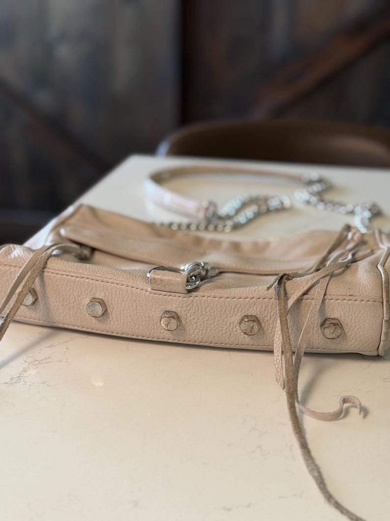 Peut inclure: Un sac &agrave; main en cuir rose clair avec des &eacute;l&eacute;ments m&eacute;talliques argent&eacute;s, dont des clous et une bandouli&egrave;re en cha&icirc;ne. Le sac a une fermeture &agrave; rabat et des franges. Le sac est pos&eacute; sur une surface blanche.