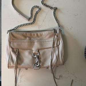 Peut inclure: Sac bandouli&egrave;re en cuir beige clair avec une cha&icirc;ne argent&eacute;e. Le sac a une poche avant &agrave; rabat, une fermeture &eacute;clair et de longues franges en cuir. Le sac mesure environ 25 cm de large.