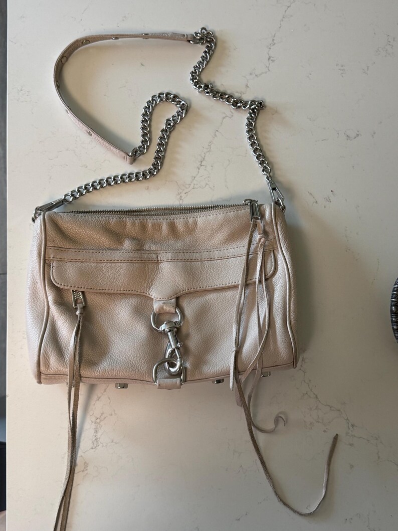 Peut inclure: Sac bandouli&egrave;re en cuir beige clair avec une cha&icirc;ne argent&eacute;e. Le sac a une poche avant &agrave; rabat, une fermeture &eacute;clair et de longues franges en cuir. Le sac mesure environ 25 cm de large.