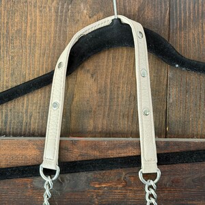 Peut inclure: Bandouli&egrave;re de sac &agrave; main cr&egrave;me avec cha&icirc;ne et accessoires argent&eacute;s. La bandouli&egrave;re est fix&eacute;e &agrave; une cha&icirc;ne argent&eacute;e. La bandouli&egrave;re est fix&eacute;e avec des rivets argent&eacute;s. Le sac est suspendu &agrave; un cintre noir sur un fond en bois.