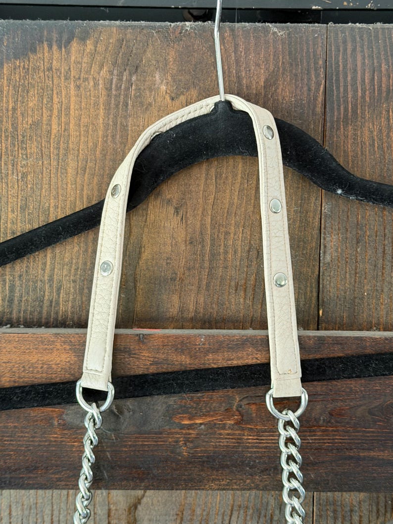 Peut inclure: Bandouli&egrave;re de sac &agrave; main cr&egrave;me avec cha&icirc;ne et accessoires argent&eacute;s. La bandouli&egrave;re est fix&eacute;e &agrave; une cha&icirc;ne argent&eacute;e. La bandouli&egrave;re est fix&eacute;e avec des rivets argent&eacute;s. Le sac est suspendu &agrave; un cintre noir sur un fond en bois.