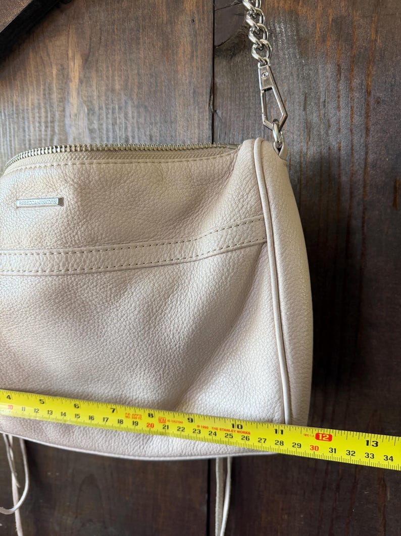 Peut inclure: Un sac &agrave; bandouli&egrave;re en cuir beige clair avec une cha&icirc;ne argent&eacute;e. Le sac a une fermeture &eacute;clair et un petit logo rectangulaire. Le sac mesure environ 30 cm de long.