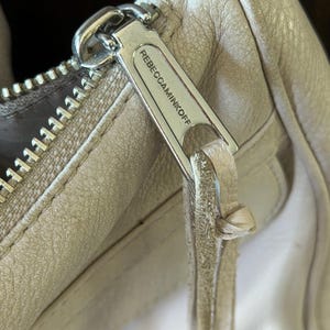 Peut inclure: Gros plan sur la tirette d'un sac &agrave; main en cuir beige clair. La tirette argent&eacute;e porte le nom de la marque "REBECCAMINKOFF". Le cuir montre des signes d'usure.