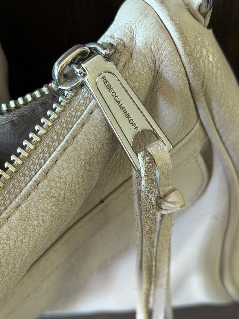 Peut inclure: Gros plan sur la tirette d'un sac &agrave; main en cuir beige clair. La tirette argent&eacute;e porte le nom de la marque "REBECCAMINKOFF". Le cuir montre des signes d'usure.