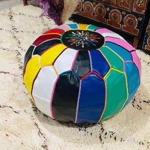 Puede incluir: Un puf redondo de cuero multicolor con un patrón geométrico. El puf presenta una variedad de colores, incluyendo amarillo, blanco, negro, azul, verde y rosa. Un parche ovalado en la parte superior tiene un diseño bordado colorido. El puf está sobre una alfombra blanca.