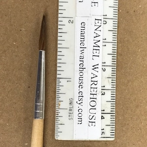 May include: A close-up of a wooden-handled paint brush with a silver ferrule and a fine point. The brush is about 1.5 inches long and is measured against a ruler.