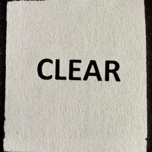 Op de afbeelding: Een witte deurmat met een zwarte rand en het woord "CLEAR" in het zwart gedrukt.