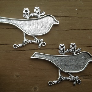 Könnte beinhalten: Zwei silberne Vogel-Anhänger aus Metall mit einem Blumendesign auf der Rückseite. Die Vögel sitzen auf einem Ast mit einer Schlaufe zum Aufhängen.