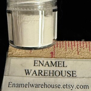 Op de afbeelding: Een klein, helder glazen potje gevuld met een fijn, gebroken wit poeder. Het potje staat op een houten liniaal met rode markeringen. De tekst "ENAMEL WAREHOUSE" en de Etsy shop URL zijn zichtbaar.