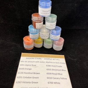 May include: A sample set of Mason Stain paint colors in small jars. The colors include Alpine Rose, Orange, Hazelnut Brown, Celadon Green, Victoria Green, Vivid Purple, Zirconium Blue, Royal Blue, Canary Yellow, and White. The jars are arranged in a pyramid shape on a box with the text "MASON STAINS SAMPLE SET #1" and "www.enamel-warehouse.com".